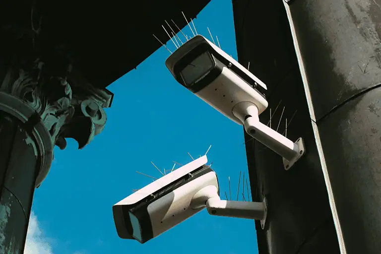 Two CCTV surveillance cameras mounted above and photographed from below against a blue sky, each pointing in a different direction, illustrating the data monitoring reach of corporate-owned music distribution platforms.