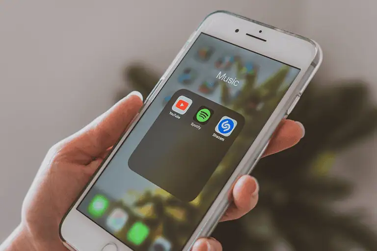 Close up of a hand holding a smartphone showing music streaming apps including Spotify, the point of contact between a listener and the platforms